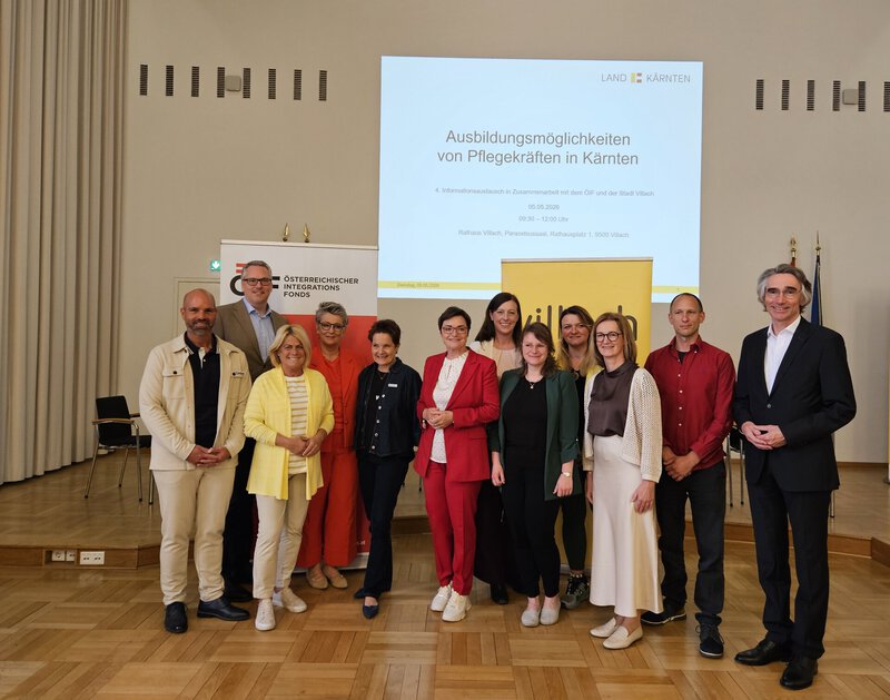 Eine Gruppe von Personen, darunter Männer und Frauen, steht zusammen in einem Raum. Im Hintergrund ist eine Präsentation zu sehen mit dem Titel "Ausbildungsmöglichkeiten von Pflegekräften in Kärnten". Die Personen wirken engagiert und an einem Austausch interessiert.