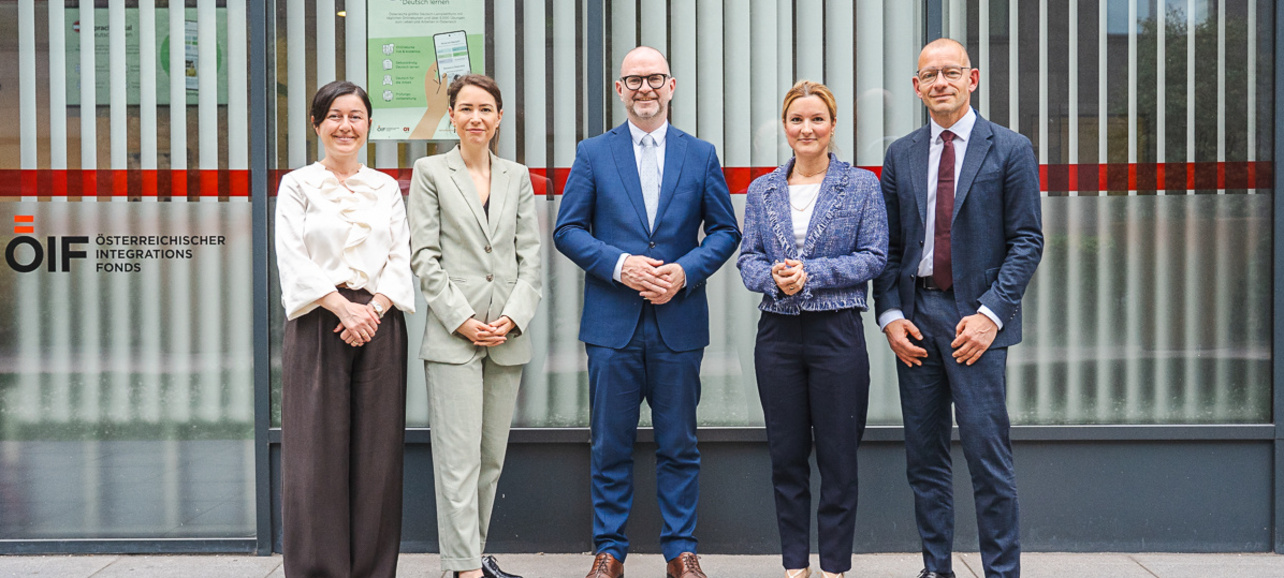 Eine Gruppe von fünf Personen steht vor einem Gebäude mit horizontalen Fensterjalousien. Sie tragen formelle Kleidung und wirken freundlich. Im Hintergrund sind Informationsplakate sichtbar.