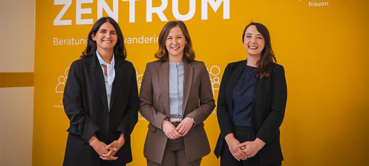 Drei Frauen stehen vor einer gelben Wand mit der Aufschrift "FRAUEN ZENTRUM". Sie tragen unterschiedliche formelle Kleidung und lächeln in die Kamera. Im Hintergrund sind Informationen über das Zentrum zu sehen.