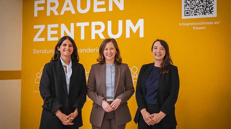 Drei Frauen stehen vor einer gelben Wand mit der Aufschrift "FRAUEN ZENTRUM". Sie tragen unterschiedliche formelle Kleidung und lächeln in die Kamera. Im Hintergrund sind Informationen über das Zentrum zu sehen.