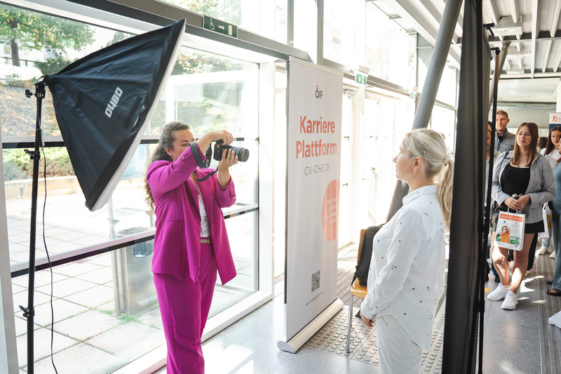 Eine Person in Pink fotografiert eine Frau in Weiß vor einem Banner zur Karriereplattform.
