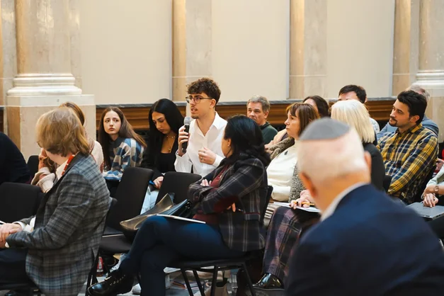 Eine Gruppe von Menschen sitzt in einem Raum, während ein junger Mann mit lockigem Haar und Brille eine Frage stellt. Die Anwesenden hören aufmerksam zu, einige schauen ihn direkt an. Der Raum hat hohe Wände mit dekorativen Elementen.