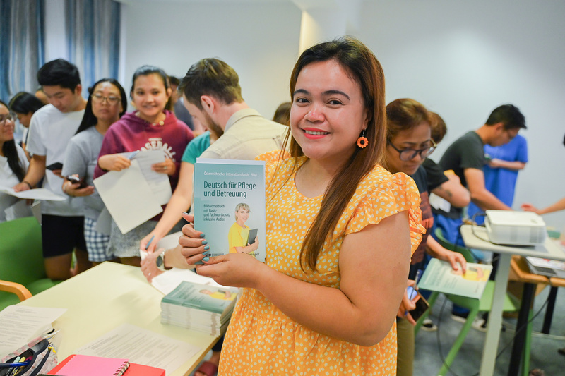 Eine lächelnde Frau hält ein Buch mit dem Titel "Deutsch für Pflege und Betreuung" in einer Warteschlange.