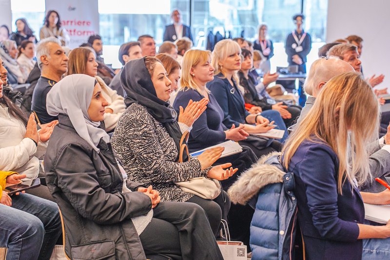 Eine Gruppe von Menschen sitzt in einem Seminarraum, einige klatschen. Die Zuschauer sind unterschiedlichen Alters und Geschlechts, tragen vielfältige Kleidung, einschließlich Kopftüchern. Im Hintergrund sind weitere Personen und ein Banner sichtbar. Die Atmosphäre scheint konzentriert und aufmerksam zu sein.
