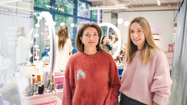 Zwei Frauen stehen lächelnd in einem Schönheitssalon, umgeben von Kosmetikprodukten und Spiegeln.