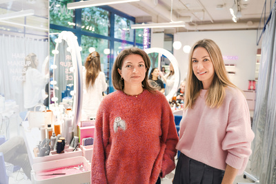 Zwei Frauen stehen lächelnd in einem Schönheitssalon, umgeben von Kosmetikprodukten und Spiegeln.