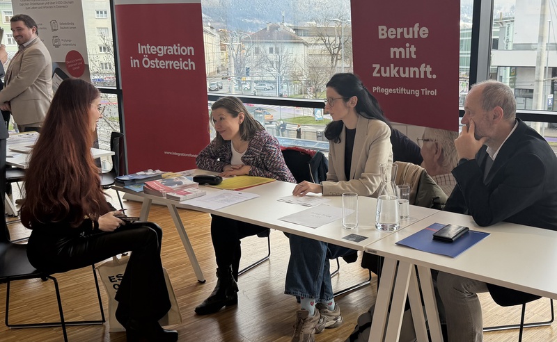 Zwei Frauen und ein Mann sitzen an einem Tisch, umgeben von Informationsmaterialien. Die Frauen diskutieren mit einer Person, die gegenüber sitzt. Im Hintergrund sind Werbeplakate für Integration in Österreich und Berufe in der Pflege zu sehen.