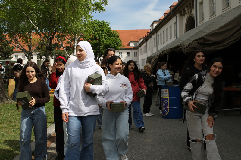 Eine Gruppe von Mädchen und jungen Frauen trägt kleine Boxen und geht fröhlich durch einen Außenbereich. Sie sind in lässiger Kleidung gekleidet und scheinen eine Veranstaltung oder einen Markt zu besuchen. Im Hintergrund sind Bäume und ein Zelt sichtbar.
