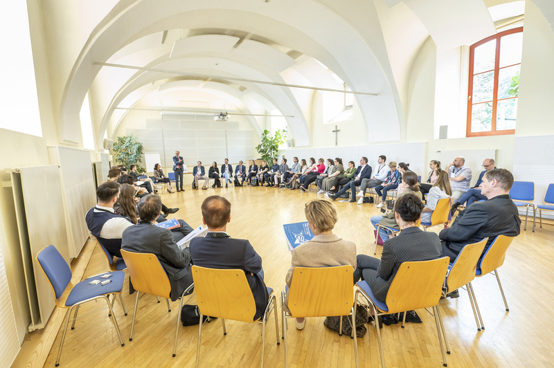 Eine Gruppe von Menschen sitzt im Kreis und hört einem Vortragenden in einem hellen Raum zu.