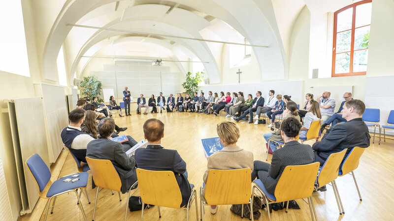 Eine Gruppe von Menschen sitzt im Kreis und hört einem Vortragenden in einem hellen Raum zu.