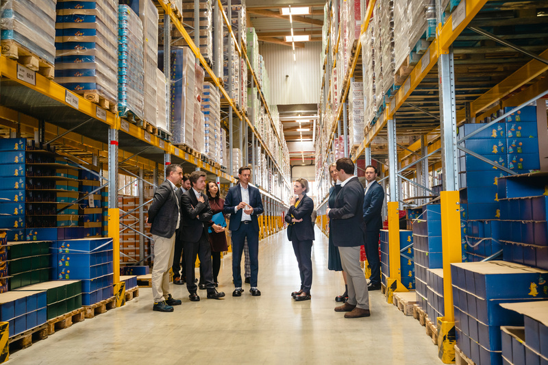 Eine Gruppe von Leuten diskutiert in einem Lagerhaus zwischen hohen Regalen mit Kisten und Produkten.