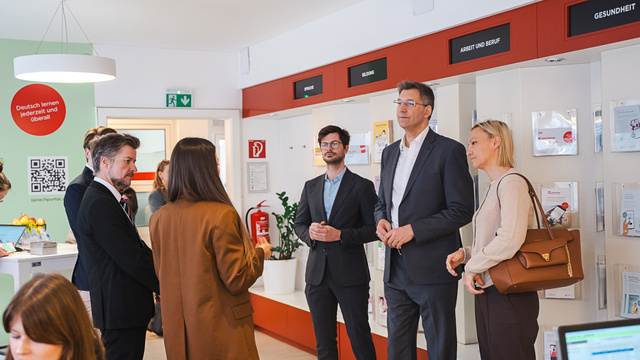 Eine Gruppe von fünf Personen steht in einem modernen Raum mit Informationswänden. Sie diskutieren miteinander, während im Hintergrund ein Schreibtisch und Informationsmaterialien sichtbar sind. Der Raum scheint ein Büro oder Beratungszentrum zu sein.