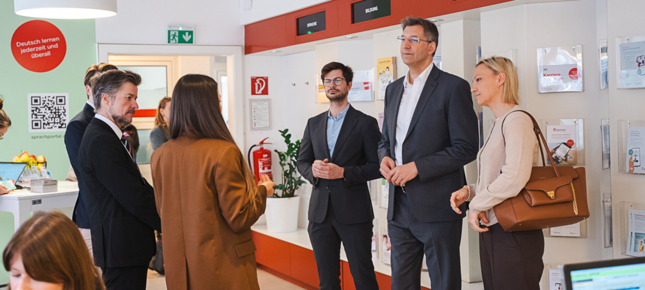 Eine Gruppe von fünf Personen steht in einem modernen Raum mit Informationswänden. Sie diskutieren miteinander, während im Hintergrund ein Schreibtisch und Informationsmaterialien sichtbar sind. Der Raum scheint ein Büro oder Beratungszentrum zu sein.