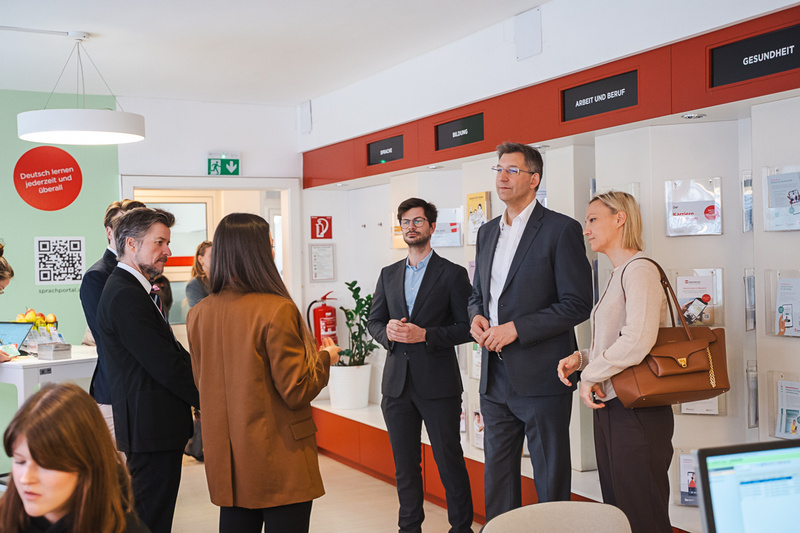 Eine Gruppe von fünf Personen steht in einem modernen Raum mit Informationswänden. Sie diskutieren miteinander, während im Hintergrund ein Schreibtisch und Informationsmaterialien sichtbar sind. Der Raum scheint ein Büro oder Beratungszentrum zu sein.