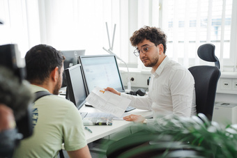 Zwei Männer in einem Büro führen ein Gespräch, einer hält Dokumente in der Hand.