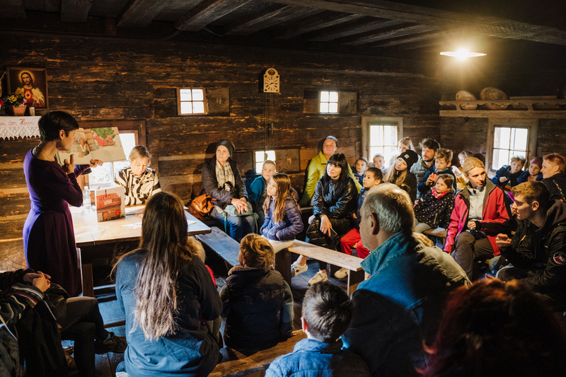 Eine Person hält ein Buch in einem Holzraum, während eine Gruppe Menschen aufmerksam zuhört.