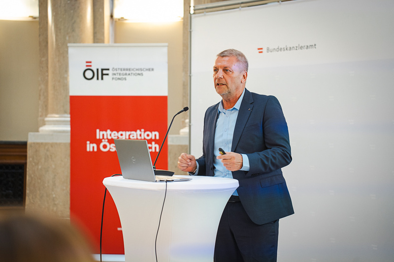 Ein Mann steht an einem Podium und spricht. Neben ihm steht ein Laptop und im Hintergrund ist ein Banner mit der Aufschrift „Integration in Österreich“ sowie das Logo des Integrationsfonds zu sehen. Das Setting scheint eine offizielle Veranstaltung zu sein.