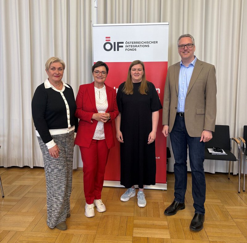 Vier Personen stehen gemeinsam vor einem Banner des Österreichischen Integrationsfonds. Die Frauen tragen verschiedene Outfits, eine in Rot, die anderen in Schwarz und Grau. Der Mann in einem hellen Sakko steht rechts. Der Hintergrund ist hell und schlicht.