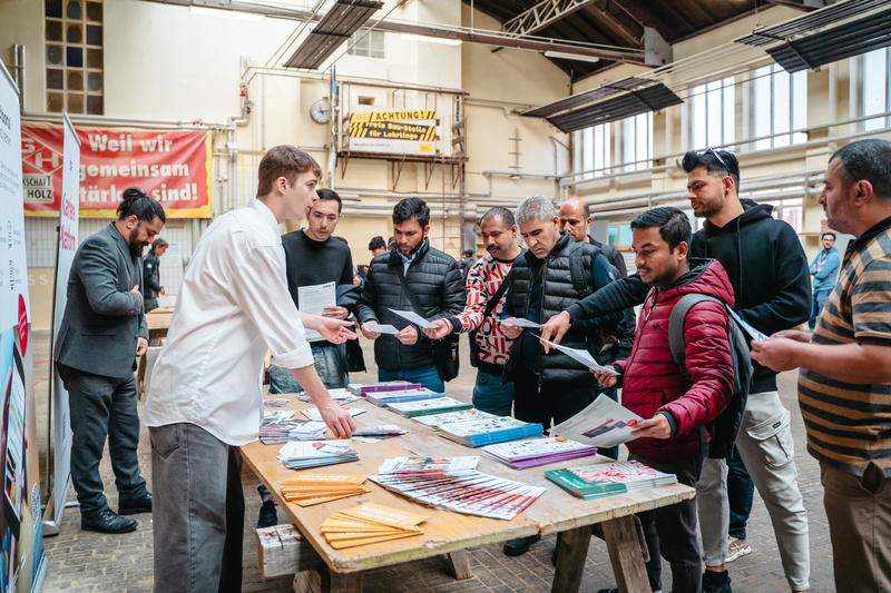 Eine Person verteilt Informationsmaterial an eine Gruppe von Menschen an einem Tisch in einer hellen Halle.