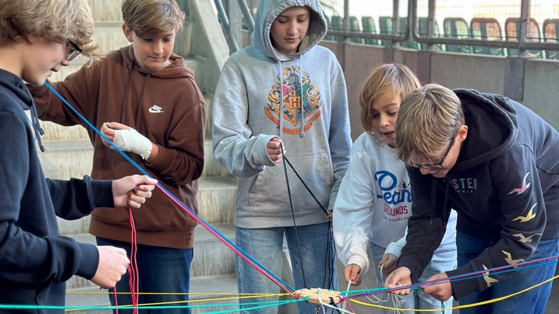 Sechs Kinder arbeiten zusammen an einem Projekt mit bunten Schnüren und einem Holzstück.