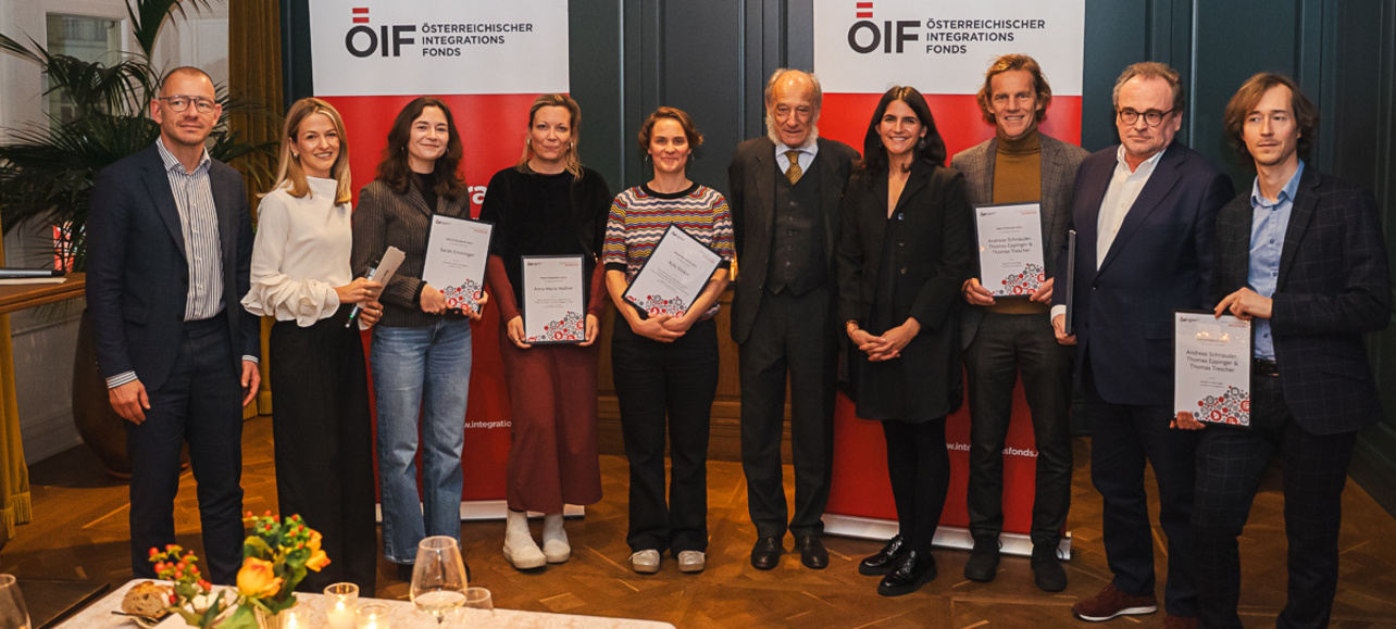 Gruppenszene mit mehreren Personen, die Urkunden in der Hand halten, vor Hintergrund mit OIF-Logo.