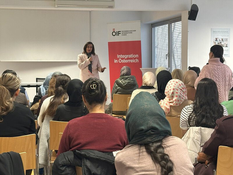 Eine Person spricht vor einer Gruppe von Menschen in einem Seminarraum. Im Hintergrund steht ein Banner mit dem Schriftzug "Integration in Österreich". Die Anwesenden tragen verschiedene Kopfbedeckungen und hören aufmerksam zu.