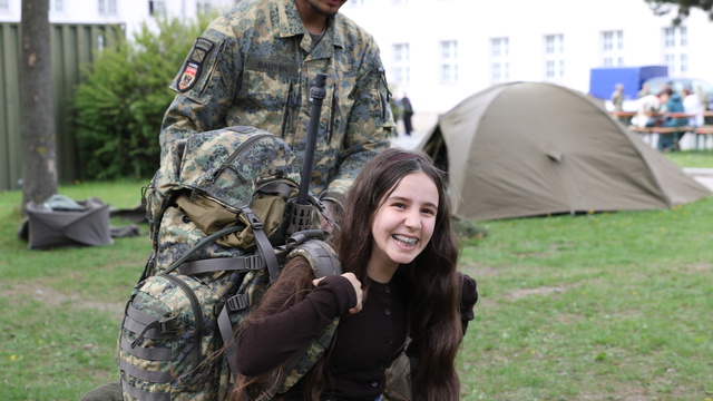 Ein Soldat hilft einem lächelnden Mädchen, einen schweren Rucksack zu tragen. Im Hintergrund sind Zelte zu sehen und es herrscht eine grüne Wiese.
