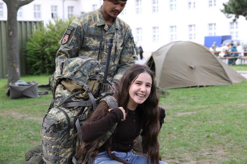 Ein Soldat hilft einem lächelnden Mädchen, einen schweren Rucksack zu tragen. Im Hintergrund sind Zelte zu sehen und es herrscht eine grüne Wiese.