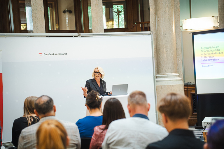 Eine Frau steht an einem Podium und präsentiert während einer Veranstaltung im Bundeskanzleramt. Sie hat einen Laptop vor sich und spricht zu einem Publikum, das ihr aufmerksam zuhört. Im Hintergrund sind lichtdurchflutete Fenster und eine Präsentation auf einem Bildschirm zu sehen.