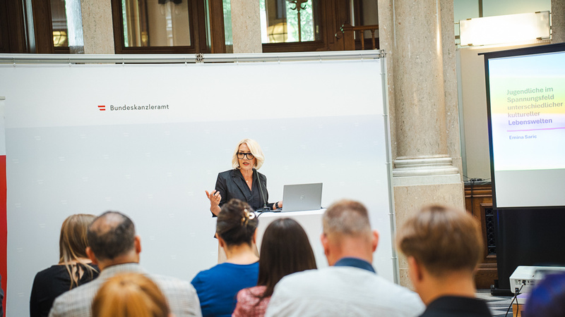 Eine Frau steht an einem Podium und präsentiert während einer Veranstaltung im Bundeskanzleramt. Sie hat einen Laptop vor sich und spricht zu einem Publikum, das ihr aufmerksam zuhört. Im Hintergrund sind lichtdurchflutete Fenster und eine Präsentation auf einem Bildschirm zu sehen.