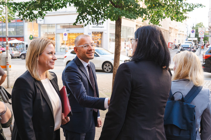 Vier Personen stehen an einer Straße und schütteln sich die Hände. Bäume und Geschäfte im Hintergrund sind sichtbar.