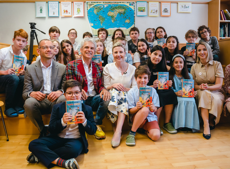 Eine Gruppe von Kindern und Erwachsenen hält Bücher in die Kamera und lächelt zusammen.