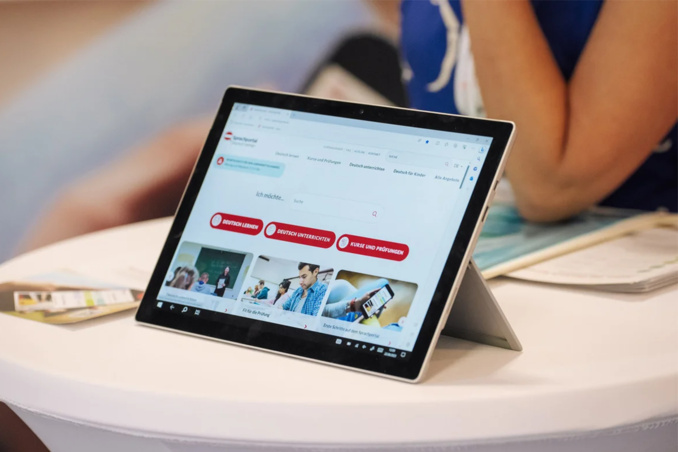 Ein Tablet steht auf einem Tisch und zeigt eine Webseite mit mehreren Bildern und roten Schaltflächen. Im Hintergrund ist eine Person zu sehen, die Teil eines Gesprächs ist. Der Fokus liegt auf dem Tablet und den dargestellten Inhalten.