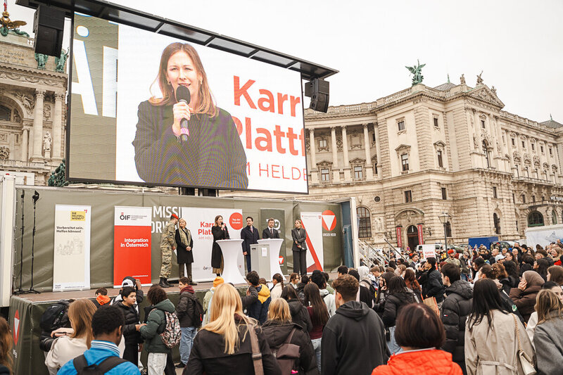 Eine Frau hält eine Rede vor einer großen Menschenmenge auf einem öffentlichen Platz.