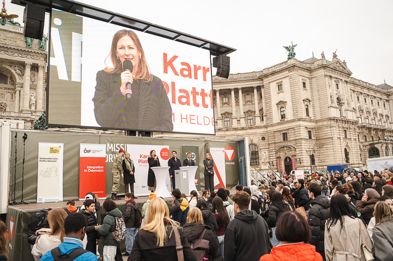 Eine Frau hält eine Rede vor einer großen Menschenmenge auf einem öffentlichen Platz.