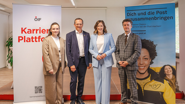Eine Gruppe von vier Personen steht vor zwei Bannern in einem Raum. Links ist ein Banner mit der Aufschrift „Karrieren Plattform“. Rechts steht ein Banner mit dem Text „Dich und die Post zusammenbringen“. Alle Personen tragen formelle Kleidung und lächeln in die Kamera.