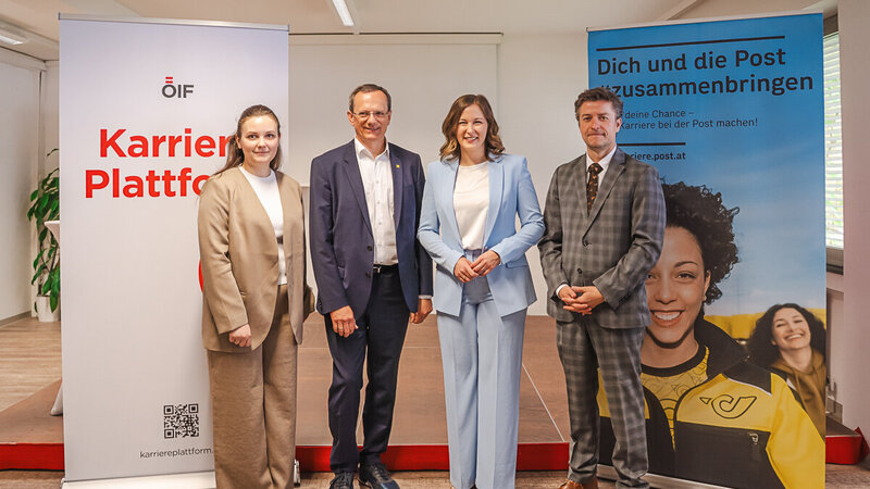 Eine Gruppe von vier Personen steht vor zwei Bannern in einem Raum. Links ist ein Banner mit der Aufschrift „Karrieren Plattform“. Rechts steht ein Banner mit dem Text „Dich und die Post zusammenbringen“. Alle Personen tragen formelle Kleidung und lächeln in die Kamera.