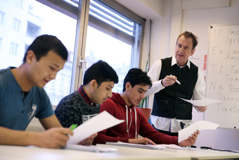 Im Bild sind vier Personen in einem Klassenzimmer zu sehen. Drei Schüler sitzen am Tisch und schauen auf ihre Unterlagen, während ein Lehrer an der Tafel steht und mit einem Stift in der Hand spricht. Tageslicht fällt durch große Fenster in den Raum.