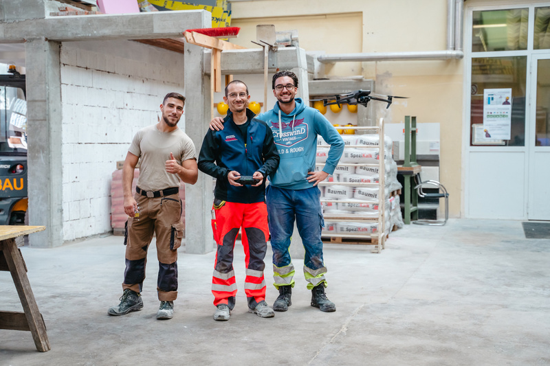 Drei Männer in Arbeitskleidung stehen in einer Baustelle und lächeln, einer hält eine Fernbedienung.