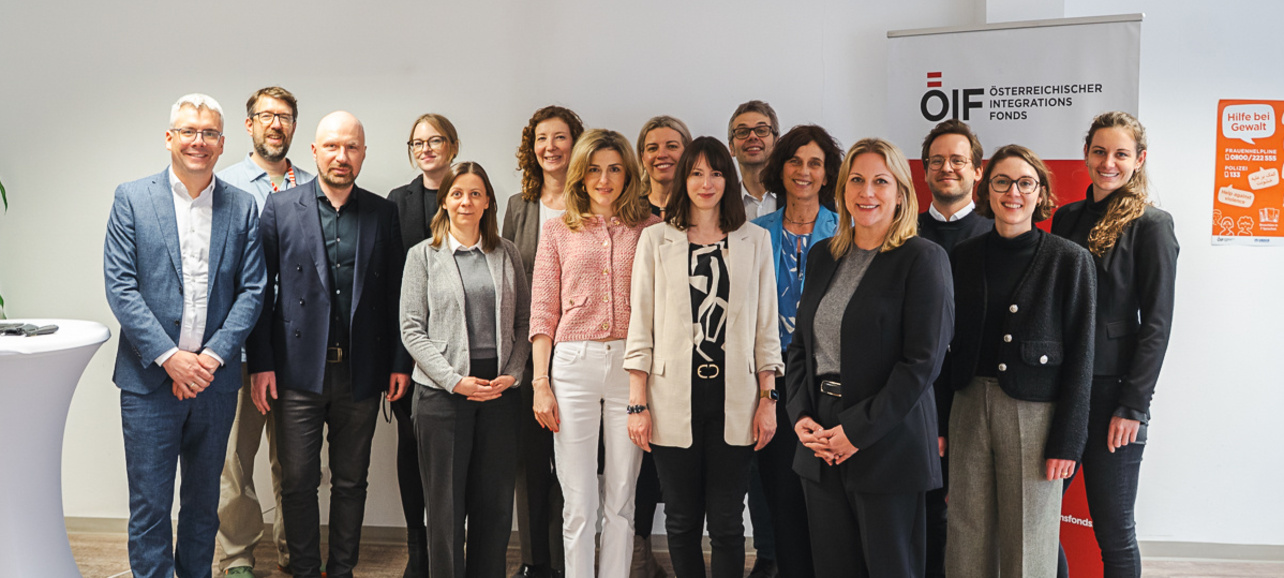Eine Gruppe von 16 Personen steht vor einem Banner des Österreichischen Austauschdienstes. Sie tragen verschiedene berufliche Kleidung und lächeln in die Kamera. Im Hintergrund ist ein heller, moderner Raum zu sehen.