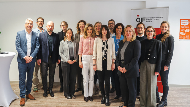 Eine Gruppe von 16 Personen steht vor einem Banner des Österreichischen Austauschdienstes. Sie tragen verschiedene berufliche Kleidung und lächeln in die Kamera. Im Hintergrund ist ein heller, moderner Raum zu sehen.
