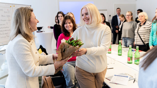 Zwei Frauen tauschen einen Blumenstrauß während eines Meetings. Teilnehmerinnen beobachten den Moment.