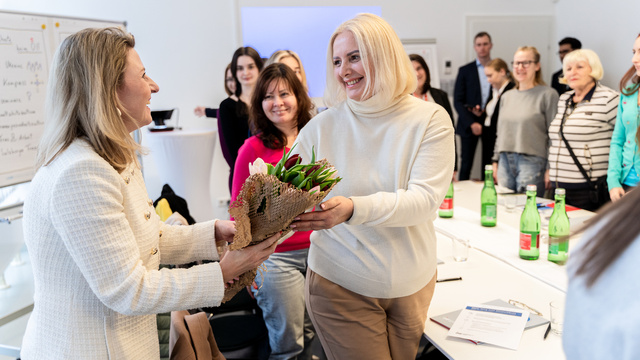 Zwei Frauen tauschen einen Blumenstrauß während eines Meetings. Teilnehmerinnen beobachten den Moment.