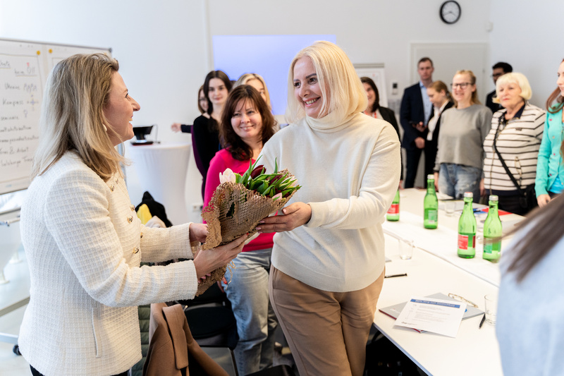 Zwei Frauen tauschen einen Blumenstrauß während eines Meetings. Teilnehmerinnen beobachten den Moment.
