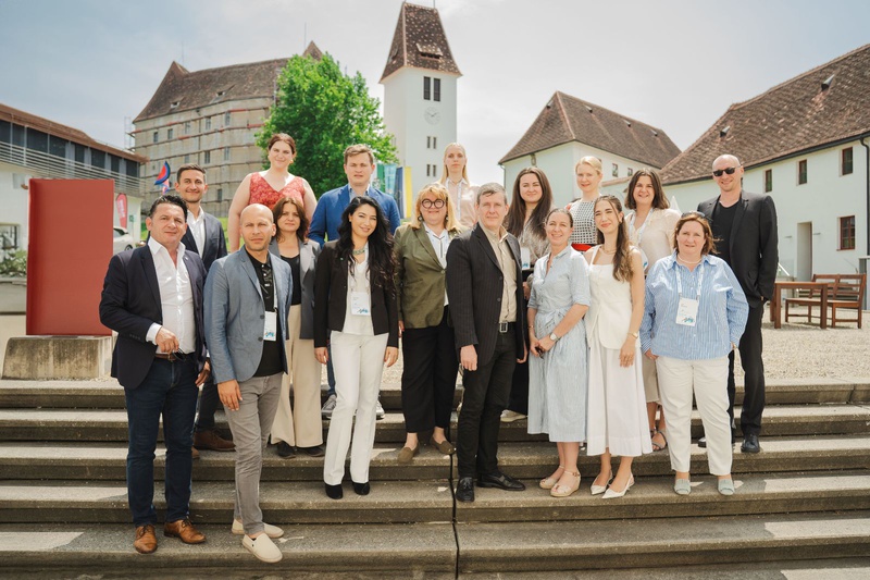 Gruppenfoto von 21 Personen auf Treppe vor einem historischen Gebäude in der Sonne.