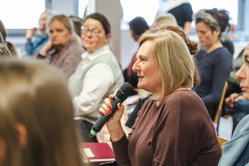 Eine Frau mit Mikrofon stellt eine Frage, während andere Personen im Hintergrund aufmerksam zuhören.