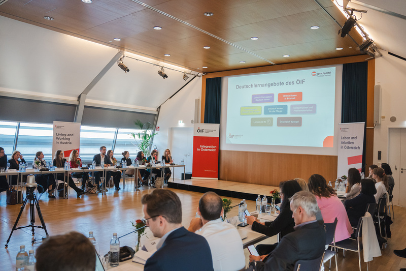 Konferenz über Deutschlernangebote in Österreich mit Teilnehmenden an einem Tisch und Präsentation im Hintergrund.