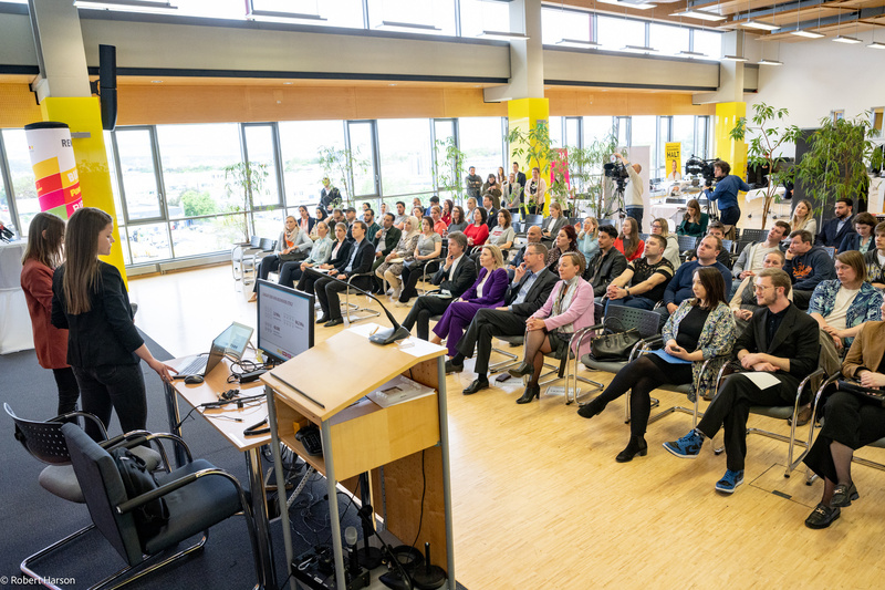 Zwei Vortragende präsentieren vor einem Publikum in einem hellen, modernen Raum mit großen Fenstern.