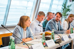 Eine Frau spricht in einem Konferenzraum, mehrere Personen sitzen am Tisch und hören aufmerksam zu.