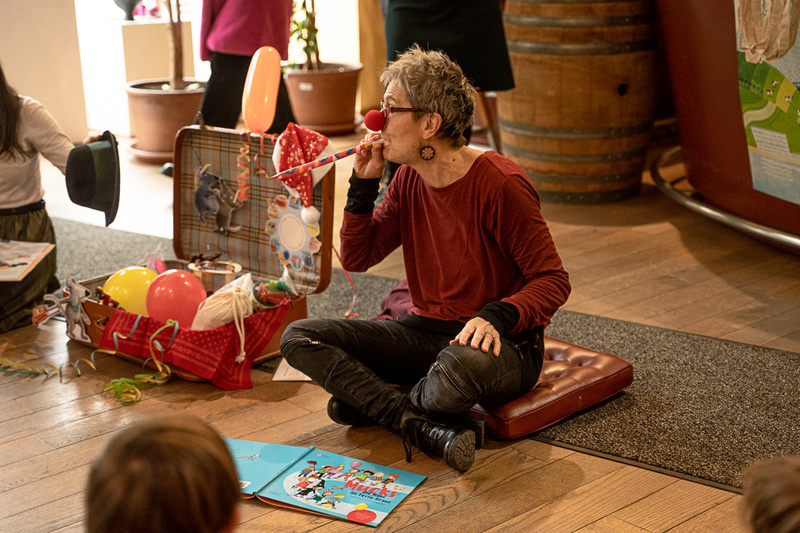 Eine Person mit einer roten Nase sitzt und spielt ein Blasinstrument, umgeben von bunten Luftballons und Spielzeug.